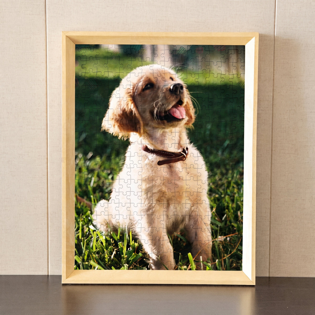 A framed custom jigsaw puzzle featuring a photo of a cute golden retriever puppy sitting on green grass, displayed on a dark surface against a beige wall.
